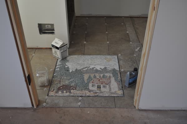 A mosaic set into the tile floor — a mountain scene with a house among pines, laid in small stones.