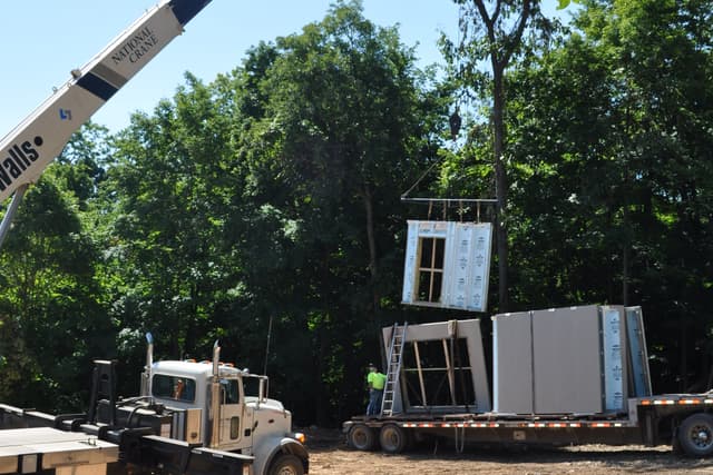 A precast wall panel swinging from a crane hook above a graded building pad, the boom truck braced below