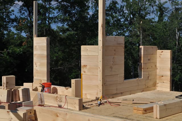 Log walls at full height with door and window openings framed by the courses, posts standing for the roof