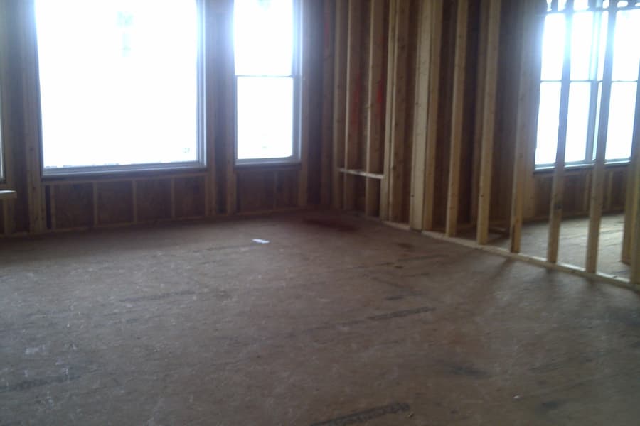A framed room with three tall window openings letting in white daylight, bare subfloor underfoot