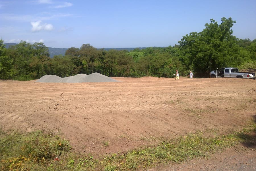 A graded building pad on a hilltop, gravel stockpiled at one end and a pickup at the other, cleared treeline and hazy ridges beyond
