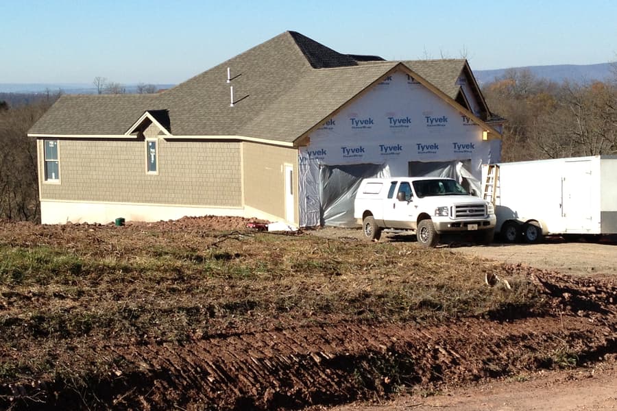 The garage end under housewrap with a truck and trailer parked at the opening, fresh earth graded around
