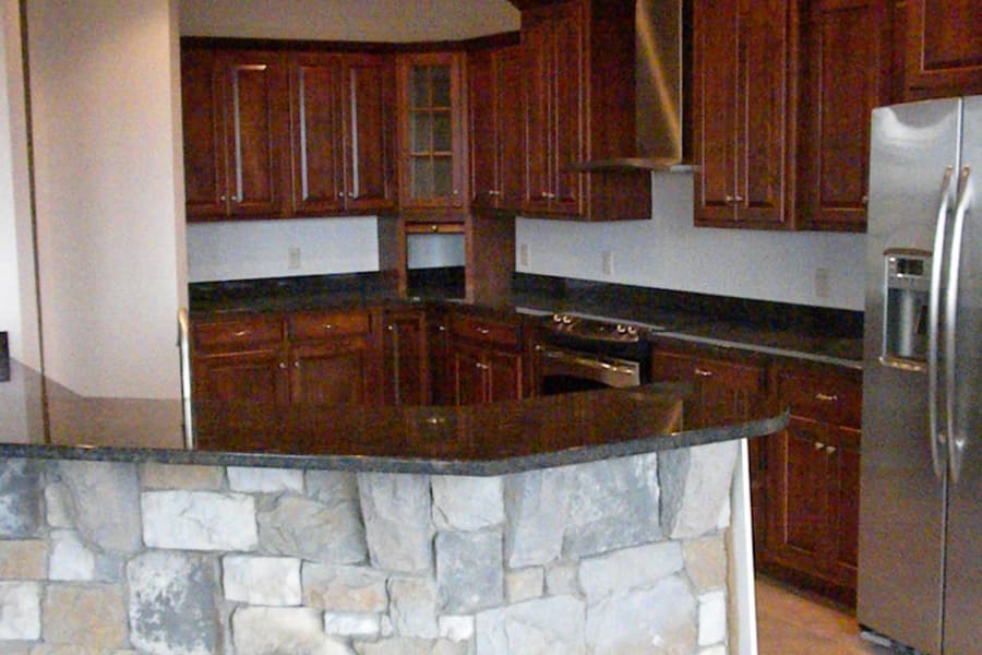 A kitchen with dark cabinets and a stone-faced island, wall oven and stainless refrigerator under recessed lights