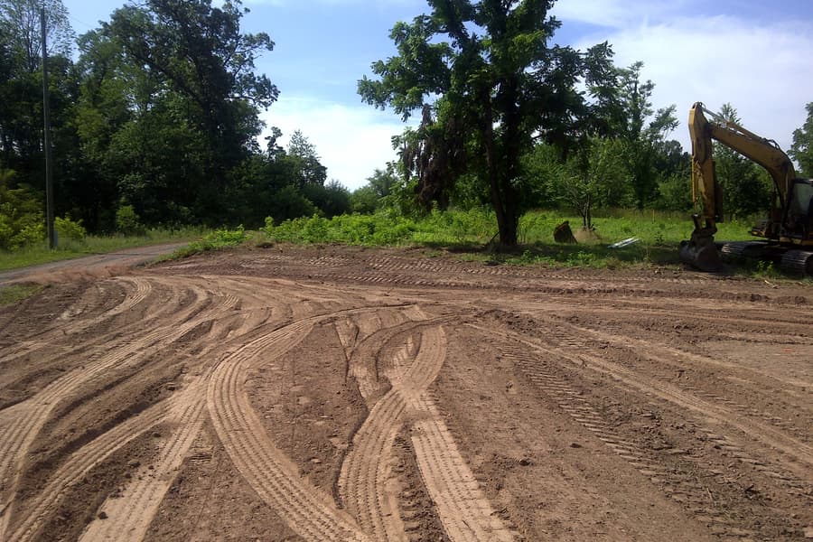 Tire-rutted earth in the foreground with one large hardwood left standing, an excavator parked beyond it