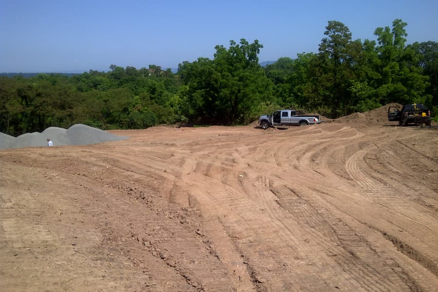 The graded pad from one corner, gravel stockpiles along the left and two vehicles parked at the treeline