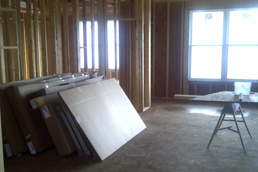 The interior framed and roofed, sheet goods stacked against a stud wall and a sawhorse standing in the open floor