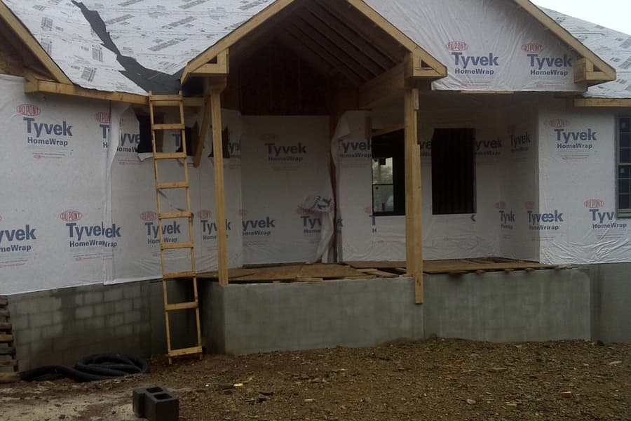 The house under housewrap with its porch gable framed, an extension ladder against the wall and block foundation below