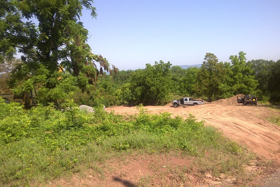 A new dirt access road cut through green scrub, a pickup and machinery parked along it, the valley opening on the right