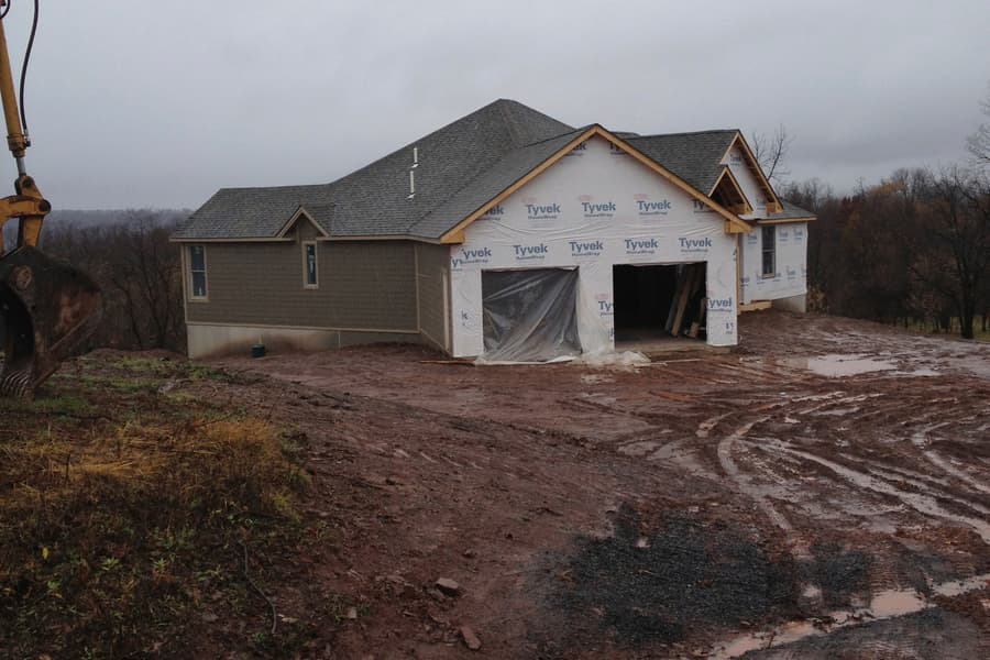 The house from the drive under housewrap, garage doors open, wet rutted ground in front