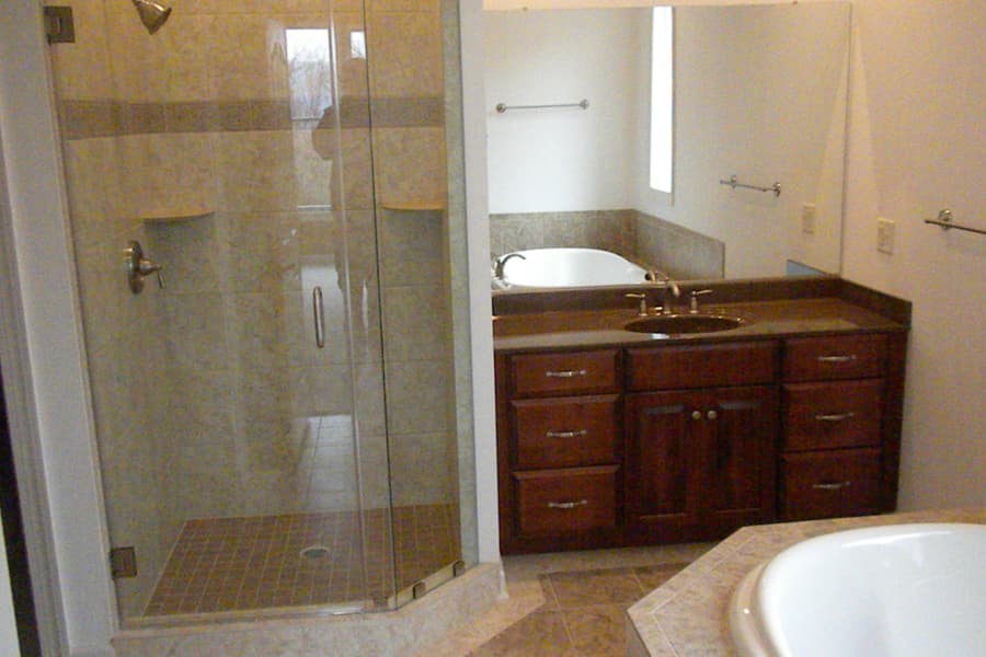 A bathroom with a glass corner shower, a tiled tub deck and a dark vanity between them